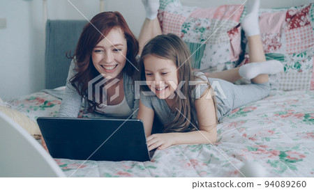 Happy cute daughter and young mother have online video call with father or grandmother using laptop computer lying on bed in cozy light bedroom at home. Happy cute daughter and young mother have online video call with father or grandmother using laptop computer lying on bed in cozy light bedroom at home. 94089260