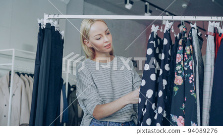 Pretty blond girl is choosing fashionable clothes in boutique. She is looking through fine garments and taking polka-dotted dress from the rails, examining its quality and length. Pretty blond girl is choosing fashionable clothes in boutique. She is looking through fine garments and taking polka-dotted dress from the rails, examining its quality and length. 94089276