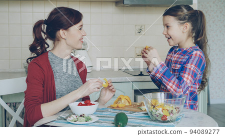Happy family of young mother and cute cheerful daughter have fun grimacing silly with vegetables while cooking in the kitchen at home 94089277