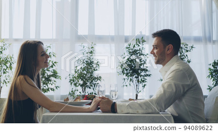 Young handsome man is talking to his girlfriend on date in fancy restaurant, drinking champagne and taking her hand with love and care. Romance and relationships concept. Young handsome man is talking to his girlfriend on date in fancy restaurant, drinking champagne and taking her hand with love and care. Romance and relationships concept. 94089622