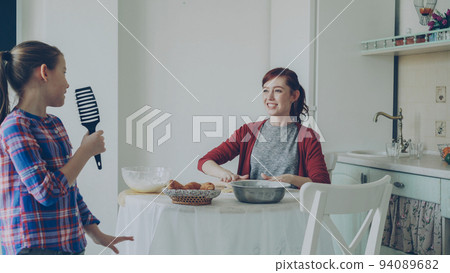 Young mother rolling dough while cooking and her little funny daughter walking around and singing with comb dancing in the kitchen on weekend. Family, food, home and people concept 94089682