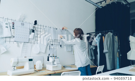 Female clothing designer is taking sketches from studio table and putting them on wall with other drawings of trendy women's garments. Creative thinking concept. 94089718