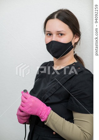 The pedicure master stands against the background of a white wall and holds an apparatus with an abrasive disc.  94092259