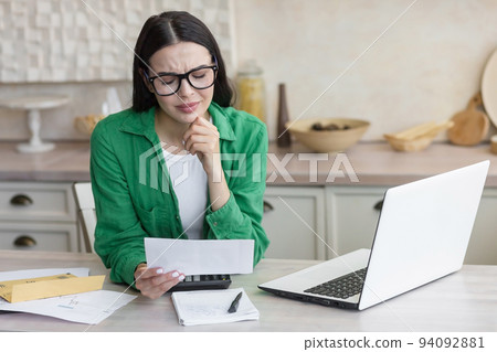 Divorce. A young beautiful woman received divorce papers. Shocked, upset. Sitting in the kitchen at home, wearing glasses and a green shirt, holding a letter in his hands. 94092881