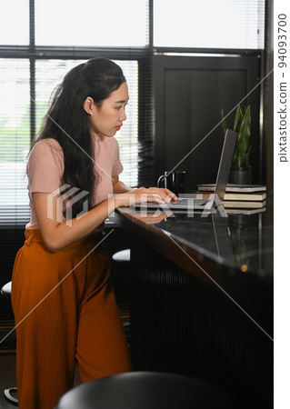 Side view of young woman working online with laptop comouter at her apartment Side view of young woman working online with laptop comouter at her apartment 94093700