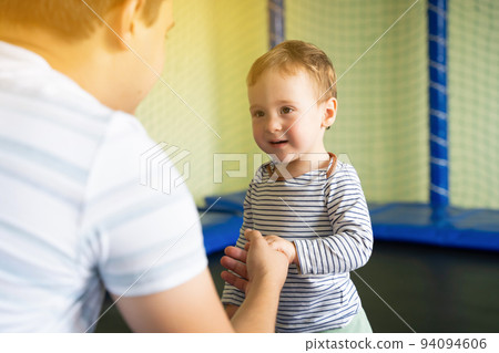 Dad holds the hand of a boy jumping on a trampoline. Family time. Boy of one and a half years. Active entertainment. Toddler. Focus on the boy's eyes Dad holds the hand of a boy jumping on a trampoline. Family time. Boy of one and a half years. Active entertainment. Toddler. Focus on the boy's eyes 94094606