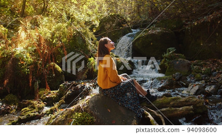 Young woman is sitting on the rock nearby the cascading water stream among the autumn forest. girl enjoys the scenic landscape of colourful nature in sunny day. caucasian mountains in fall season. Young woman is sitting on the rock nearby the cascading water stream among the autumn forest. girl enjoys the scenic landscape of colourful nature in sunny day. caucasian mountains in fall season. 94098600