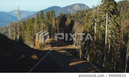 Mountain road among the deep forest in fall season. treetops painted in colorful autumn colours in the sun rays. forested mountain peaks of caucasus mountain range on the clear sky background. 94098632