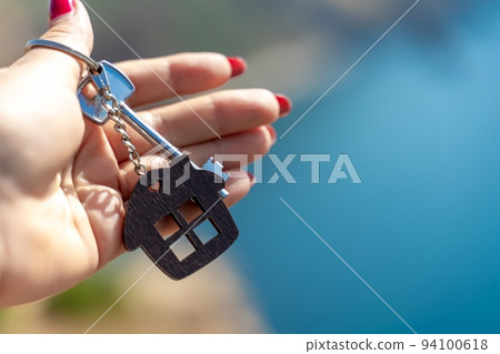 Keys with a keychain in the shape of a house in a female hand against the backdrop of the sea. The concept of buying a house, apartment, real estate, young family, mortgage loan, rental housing. Keys with a keychain in the shape of a house in a female hand against the backdrop of the sea. The concept of buying a house, apartment, real estate, young family, mortgage loan, rental housing. 94100618