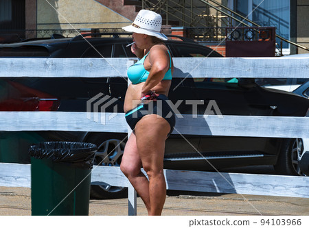 A plump woman in a swimsuit stands by the fence and smokes a cigarette. A plump woman in a swimsuit stands by the fence and smokes a cigarette. 94103966