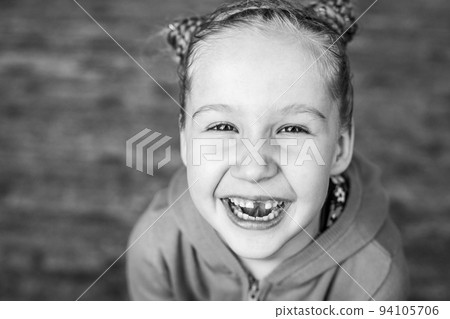 Black and white close-up portrait of a laughing little girl with a missing front tooth. Cheerful child. Real sincere emotions, happiness, childhood. Black and white close-up portrait of a laughing little girl with a missing front tooth. Cheerful child. Real sincere emotions, happiness, childhood. 94105706