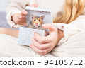 A cute fluffy golden hamster looks out of a gift box in the hands of a girl in knitted home clothes and looks at camera. Beautiful postcard with an animal theme. Pet as a gift for birthday, christmas. 94105712