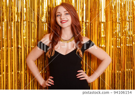 Portrait of young charming woman with red lips, long red hair smiling to camera with golden tinsels on background, keeping hand son hips, celebrating party. 94106154