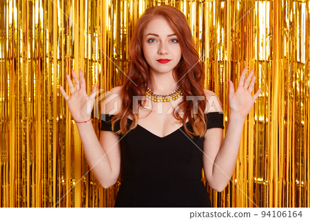 Girl being pissed and losing temper while posing against wall with golden tinsel, european red haired girl with beautiful curls, looks angry, raised hands up, being offended during party. 94106164