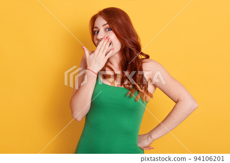 Red haired young woman, looks happy and excited, being surprised and amazed, covering mouth with hands, giggling with cute expression against yellow background. Red haired young woman, looks happy and excited, being surprised and amazed, covering mouth with hands, giggling with cute expression against yellow background. 94106201