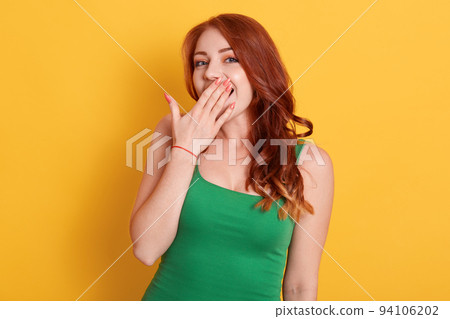 Beautiful red haired woman yawns isolated over yellow background, wears casual green t shirt, keeps palm near mouth, pleasant looking female looks at camera. Beautiful red haired woman yawns isolated over yellow background, wears casual green t shirt, keeps palm near mouth, pleasant looking female looks at camera. 94106202