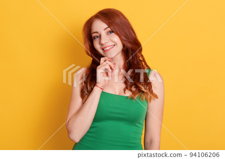 Portrait of pretty, nice looking , cute girl keeping fingers on chin, looking at camera, standing over yellow background, smiling young lady with red hair showing white teeth. Portrait of pretty, nice looking , cute girl keeping fingers on chin, looking at camera, standing over yellow background, smiling young lady with red hair showing white teeth. 94106206