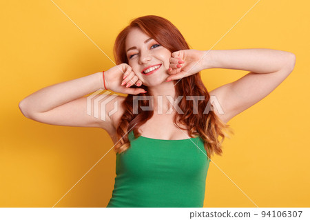 Beautiful smiling woman stretching and winking while looking directly at camera, wearing green sleeveless t shirt, red haired lady with pleasant appearance and positive emotions. Beautiful smiling woman stretching and winking while looking directly at camera, wearing green sleeveless t shirt, red haired lady with pleasant appearance and positive emotions. 94106307