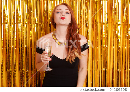 Happy woman making kiss gesture while holding glass of wine, looks at camera, wearing black dress and necklace, lady celebrating birthday or New Year. 94106308