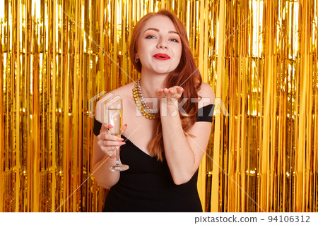 Excited pretty red haired girl holding wineglass of sparkling champagne standing isolated on sparkle background. Model sending air kisses while celebrating important event. 94106312