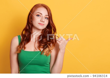 Calm woman with red hair, wears green t shirt, points with thumb, shows fantastic product, isolated over yellow background, copy space for advertisement or promo. 94106314