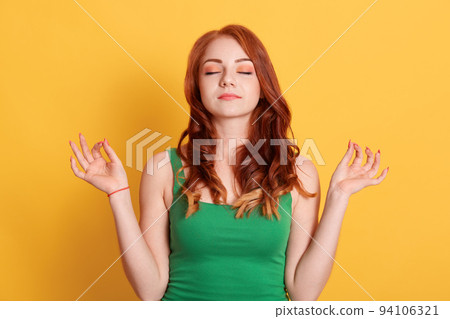 Peaceful European girl with closed eyes, practicing breathing yoga against yellow background, female wearing green t shirt, keeps eyes closed, red haired girl relaxing. 94106321