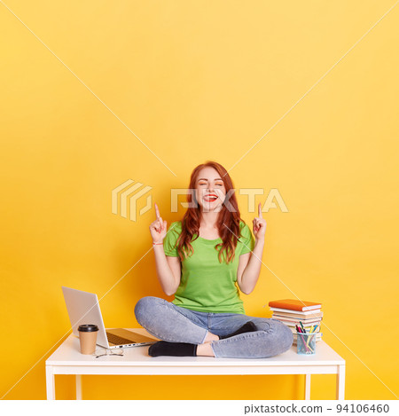 Young ginger overjoyed woman with red hair, wears jeans and green t shirt, points with both index finger upwards, shows free space for your advertising content, winking and showing tongue. Young ginger overjoyed woman with red hair, wears jeans and green t shirt, points with both index finger upwards, shows free space for your advertising content, winking and showing tongue. 94106460