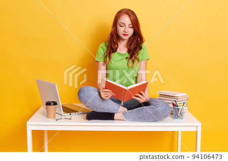 Red haired concentrated female wearing green t shirt and jeans, holding books in hands and reading, student sitting on table with crossed legs, lady surrounded with laptop, pens, books. Red haired concentrated female wearing green t shirt and jeans, holding books in hands and reading, student sitting on table with crossed legs, lady surrounded with laptop, pens, books. 94106473