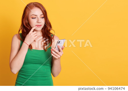 Caucasian beautiful girl using smart phone and thinking over yellow background, red haired female wearing green outfit keeping finger on her lip, looks at device screen. Caucasian beautiful girl using smart phone and thinking over yellow background, red haired female wearing green outfit keeping finger on her lip, looks at device screen. 94106474