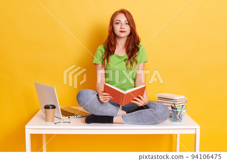 Smiling young woman with red hair, girl in green t shirt and jeans, looks at camera, posing isolated over yellow background, sits with book in hands. Smiling young woman with red hair, girl in green t shirt and jeans, looks at camera, posing isolated over yellow background, sits with book in hands. 94106475