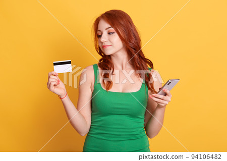 Portrait of nice attractive red haired girl holding in hands bank card and cell phone, wireless purchase, ginger girl buying things online isolated over yellow background. Portrait of nice attractive red haired girl holding in hands bank card and cell phone, wireless purchase, ginger girl buying things online isolated over yellow background. 94106482