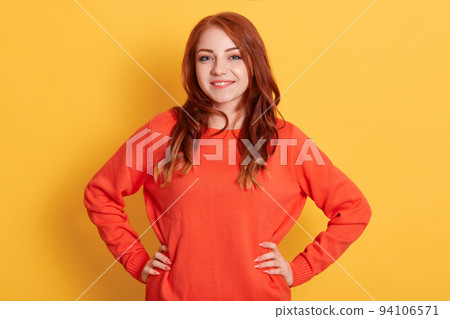 Confident young lady with red hair standing with hands on hips and looking at camera, being in good mood, smiling happily against yellow background, female wearing casual attire. Confident young lady with red hair standing with hands on hips and looking at camera, being in good mood, smiling happily against yellow background, female wearing casual attire. 94106571