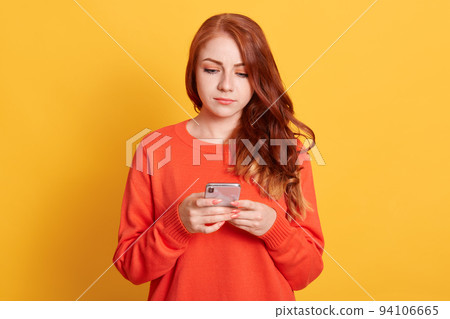 Serious young red haired woman girl in orange sweater, posing isolated on yellow wall, pensive girl using mobile phone, typing sms message or checking social networks. 94106665