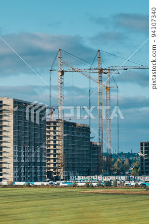 Building site background. Industrial construction cranes on background of clouds sky. Hoisting cranes, multi-storey buildings of new city districts and large green field. Project of urban area. Building site background. Industrial construction cranes on background of clouds sky. Hoisting cranes, multi-storey buildings of new city districts and large green field. Project of urban area. 94107340