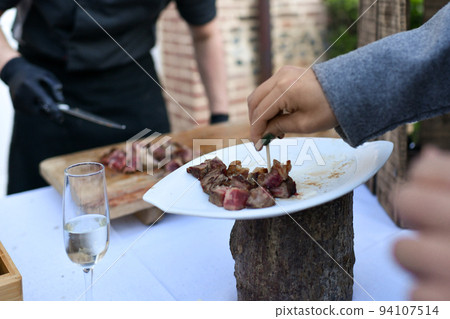 The chef cooks the veal at a plancha and the guest at the wedding tastes the meat The chef cooks the veal at a plancha and the guest at the wedding tastes the meat 94107514