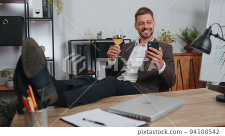 Businessman drinking champagne from glass after working, putting legs on table sitting at office 94108542