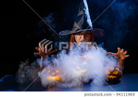 An ominous witch in a hat conjures over a jack-o-lantern. Traditional halloween characters. Mystical fog creeps over pumpkins with carved terrible faces. An evil witch casts a spell. 94111969