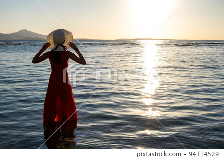 Young woman wearing long red dress and straw hat standing in sea water at the beach enjoying view of rising sun in early summer morning. 94114529