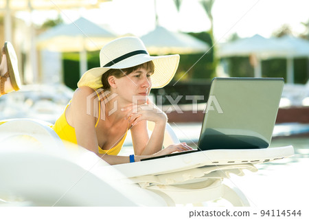 Young woman in yellow dress is laying on beach chair working on computer laptop connected to wireless internet typing text on keys in summer resort. Doing studies while travelling concept. Young woman in yellow dress is laying on beach chair working on computer laptop connected to wireless internet typing text on keys in summer resort. Doing studies while travelling concept. 94114544