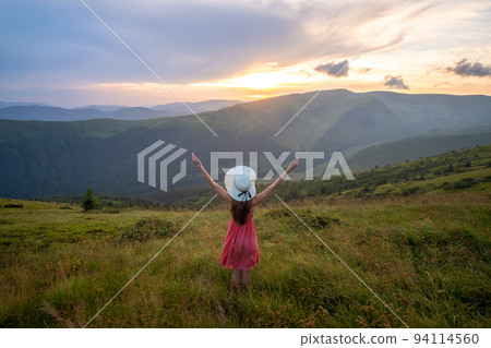 Young woman in red dress standing on grassy meadow on a windy evening in autumn mountains raising up her hands enjoying view of nature. 94114560