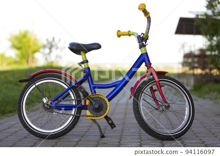 Close-up of child bright multicolored blue, yellow and red bicycle left supported on side stand in middle of empty paved street on blurred bright bokeh background. Children active lifestyle concept. Close-up of child bright multicolored blue, yellow and red bicycle left supported on side stand in middle of empty paved street on blurred bright bokeh background. Children active lifestyle concept. 94116097