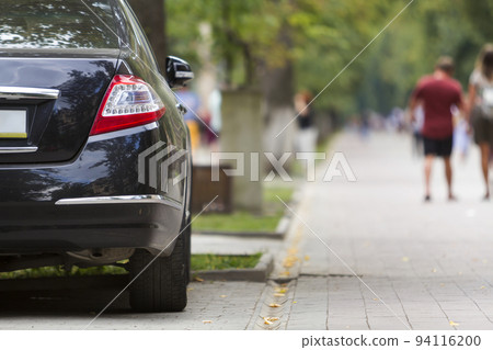 Back view part of black luxury shiny car parked on city pedestrian zone pavement on background of blurred silhouettes of people walking along green sunny summer alley. Modern lifestyle concept. Back view part of black luxury shiny car parked on city pedestrian zone pavement on background of blurred silhouettes of people walking along green sunny summer alley. Modern lifestyle concept. 94116200