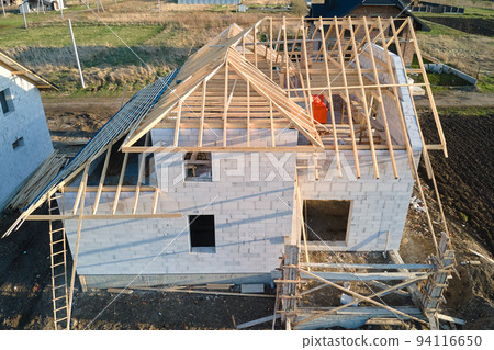 Aerial view of unfinished frame of private house with aerated lightweight concrete walls and wooden roof frame under construction. Aerial view of unfinished frame of private house with aerated lightweight concrete walls and wooden roof frame under construction. 94116650