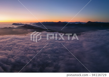 Aerial view of colorful sunrise over white dense fog with distant dark silhouettes of mountain hills on horizon Aerial view of colorful sunrise over white dense fog with distant dark silhouettes of mountain hills on horizon 94116729