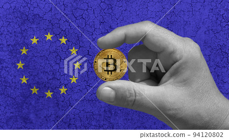 One hand hold a Bitcoin, btc, the cryptocurrency of the future. In the background the European flag on a ruined concrete wall 94120802