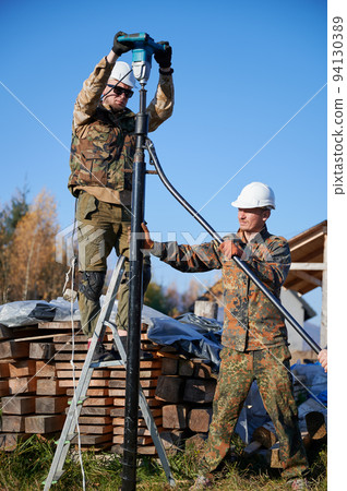 Male workers building pile foundation for wooden frame house. Men builders in white safety helmets drilling piles into the ground on blue sky background. 94130389