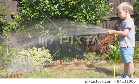 Funny little boy watering lawn plants in garden housing backyard. Adorable child playing with irrigation hose at hot sunny summer outdoors. Children help with housework. activity for kids. Childhood Funny little boy watering lawn plants in garden housing backyard. Adorable child playing with irrigation hose at hot sunny summer outdoors. Children help with housework. activity for kids. Childhood 94131000