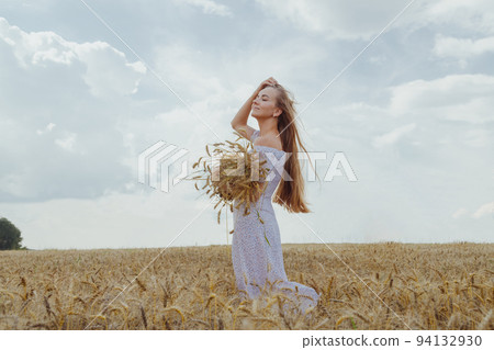 Young blonde woman with long hair stands in a wheat field with an armful of ripe ears of corn. Young blonde woman with long hair stands in a wheat field with an armful of ripe ears of corn. 94132930