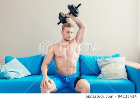 Young handsome athletic man trains with dumbbells, doing sports at home, sitting on sofa Young handsome athletic man trains with dumbbells, doing sports at home, sitting on sofa 94133434