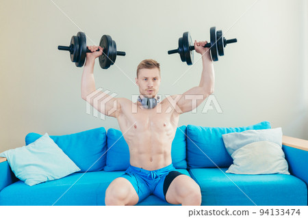 Young handsome athletic man trains with dumbbells, doing sports at home, sitting on sofa listening to music on headphones, looking at the camera Young handsome athletic man trains with dumbbells, doing sports at home, sitting on sofa listening to music on headphones, looking at the camera 94133474